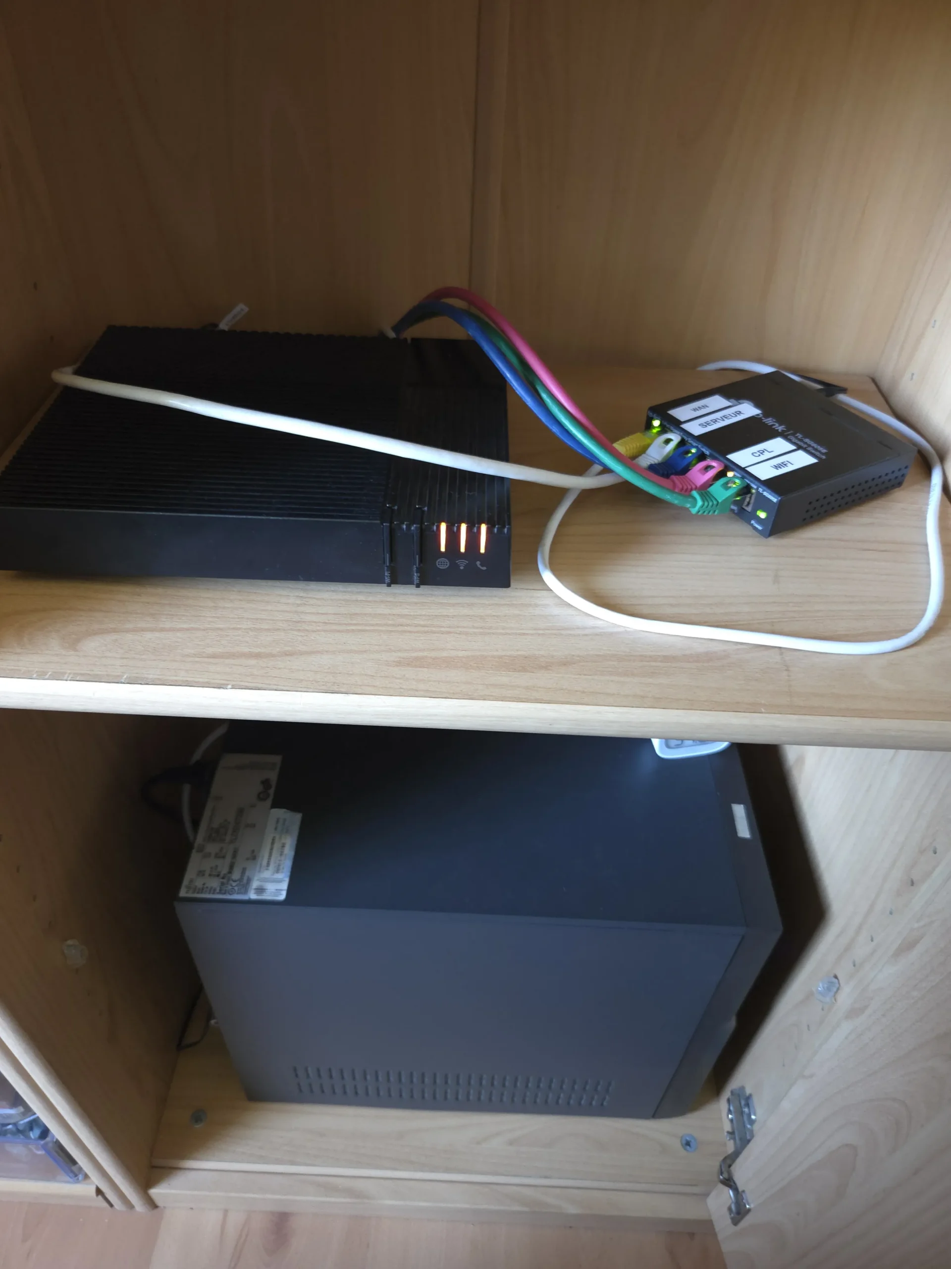 Une image représentant une box Internet avec une coque noire rectangulaire avec 3 voyants Orange à droite (en haut à gauche de l'image), un switch réseau noir avec des lumières vertes et une orange sur le port 4 qui dispose de 5 ports, tous avec un câble relié (en haut à droite) et un serveur (en bas) en avec une coque noire métallique rectangulaire (en bas). La box et le switch sont sur l'étagère du haut, tandis que le serveur et posé au fond du placard tout en bas. Le placard et l'étagère sont en bois.